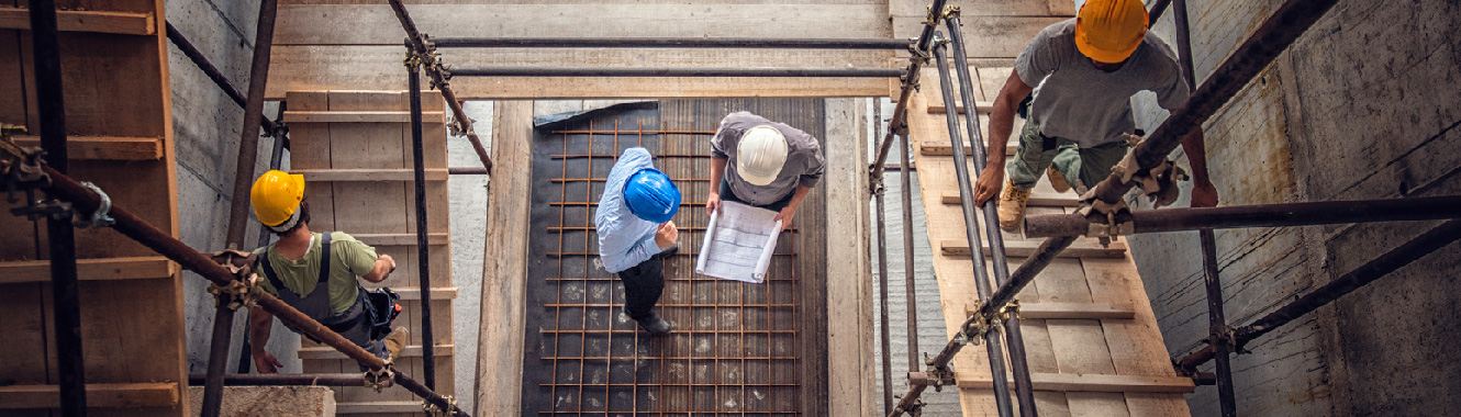 A top-down view of a construction site. Two workers talk over blueprints at the bottom of the structure. 
