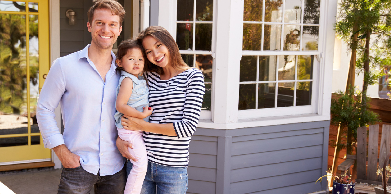A couple with their young daughter are standing outside of their newly purchased home and smiling at the camera.
